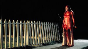 Carrie White drenched in blood, walking home at night beside a picket fence in Carrie (1976).